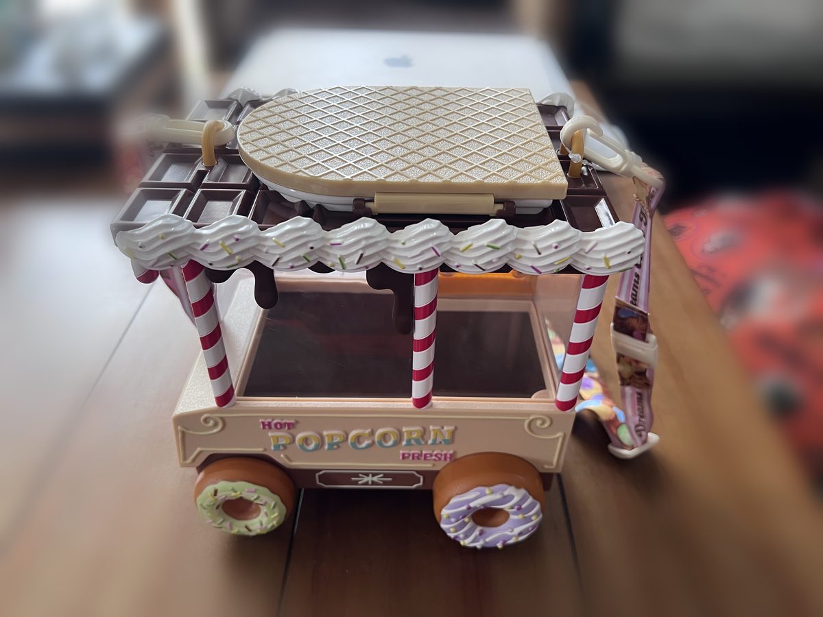 スイーツワゴン型のポップコーンバケット。チョコレートの屋根にホイップクリーム、キャンディ柄の柱、ドーナツのタイヤがついたワゴンのデザイン
